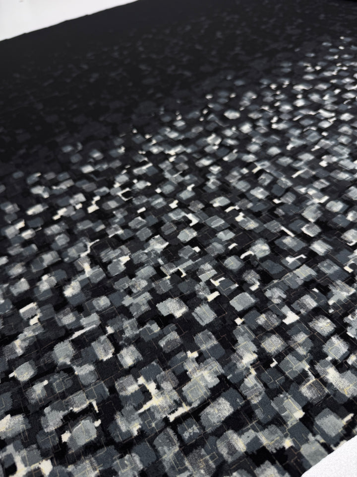 A close-up of the Designer Lycra Panel - 90cm Panel by Super Cheap Fabrics shows a textured surface with a grid of black, gray, and white brush strokes, creating an abstract pixelated effect that fades into darkness.