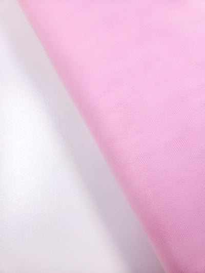 A close-up of Super Cheap Fabrics’ Soft Tulle - Baby Pink - 150cm, a gently textured fabric ideal for costumes or veils, rolled out on a light surface. Soft lighting enhances its delicate pastel appearance.