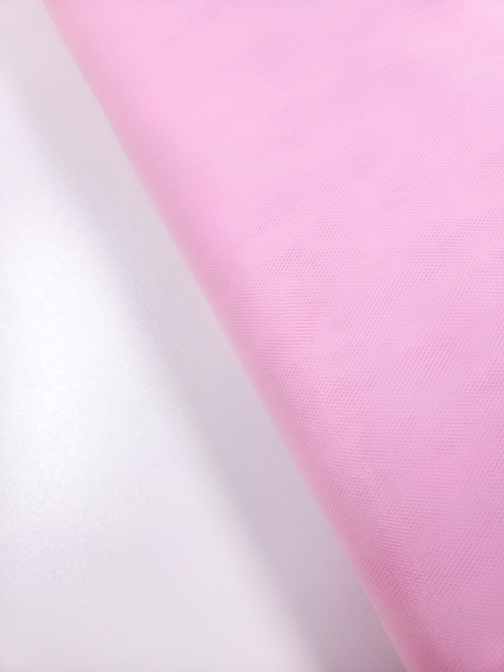 A close-up of Super Cheap Fabrics’ Soft Tulle - Baby Pink - 150cm, a gently textured fabric ideal for costumes or veils, rolled out on a light surface. Soft lighting enhances its delicate pastel appearance.