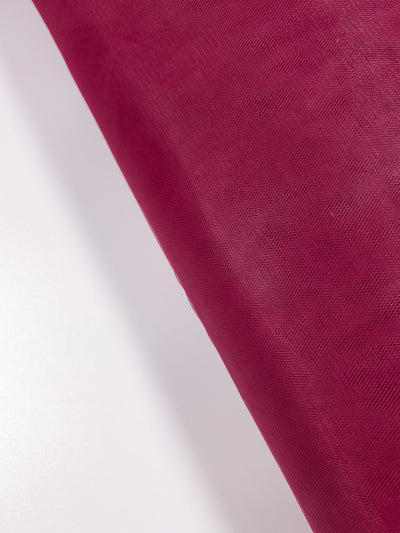 The Soft Tulle - Emboldened - 150cm by Super Cheap Fabrics, shown partially unrolled on a white surface, features fine, sheer deep red material perfect for bridal veils or costumes.