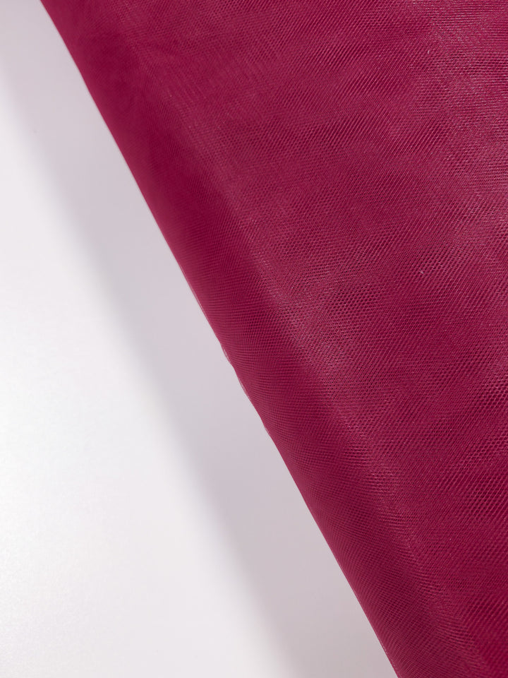 The Soft Tulle - Emboldened - 150cm by Super Cheap Fabrics, shown partially unrolled on a white surface, features fine, sheer deep red material perfect for bridal veils or costumes.