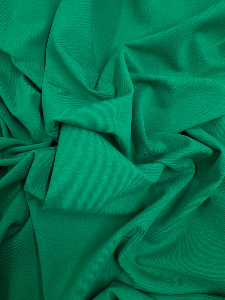 Super Cheap Fabrics’ Cotton Lycra - Deep Mint (165cm) is shown loosely gathered and folded, its green stretch fabric featuring a smooth, slightly textured surface with soft, flowing creases and gentle shadows.