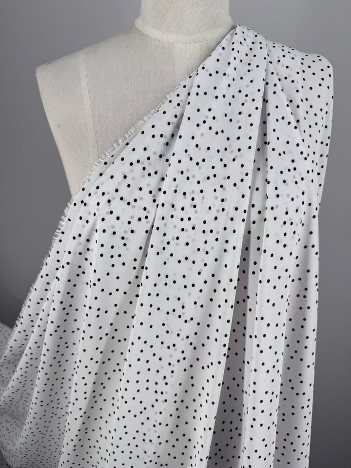 The 145cm-wide Printed Chiffon - Polka Dots fabric from Super Cheap Fabrics, featuring a white base with small black dots, elegantly drapes over a mannequin against a gray background.