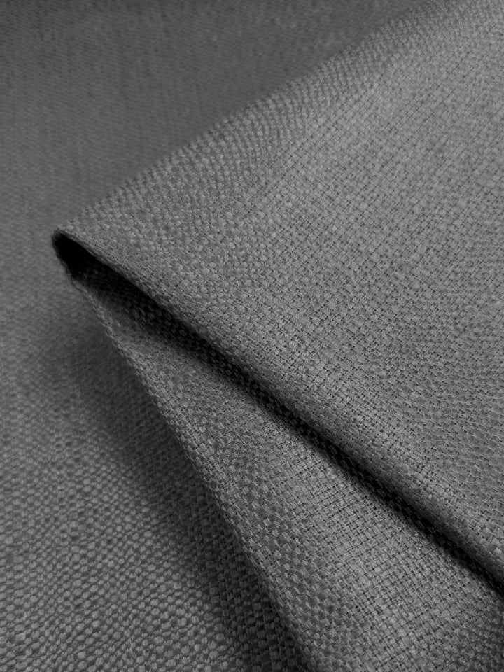 A close-up of Dorian Upholstery - Gunmetal (146cm) by Super Cheap Fabrics shows soft, textured grey fabric with a subtle woven pattern—ideal for high-end upholstery projects.