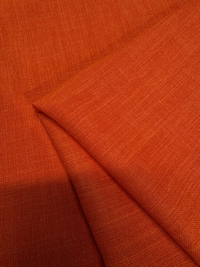 A close-up of Crepe - Puffin’s Bill - 112cm from Super Cheap Fabrics in orange, showing lightweight polyester fabric with a fine woven pattern, one piece flat and another folded—ideal for elegant evening gowns.