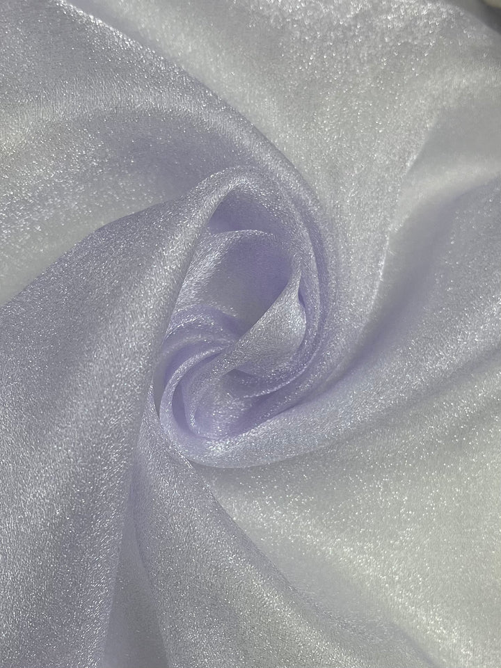 A close-up of Super Cheap Fabrics' Organza in Orchid Hush, intricately folded to create a soft, swirling pattern. This lightweight, light purple sheer material catches the light, giving it a slightly metallic, iridescent sheen.