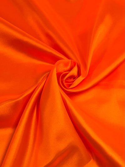 A close-up photograph showcases the smooth, vibrant orange "Satin - Orange - 150cm" fabric by Super Cheap Fabrics, artfully draped and gathered at the center to create a swirl-like pattern. The material features a shiny, satiny texture.