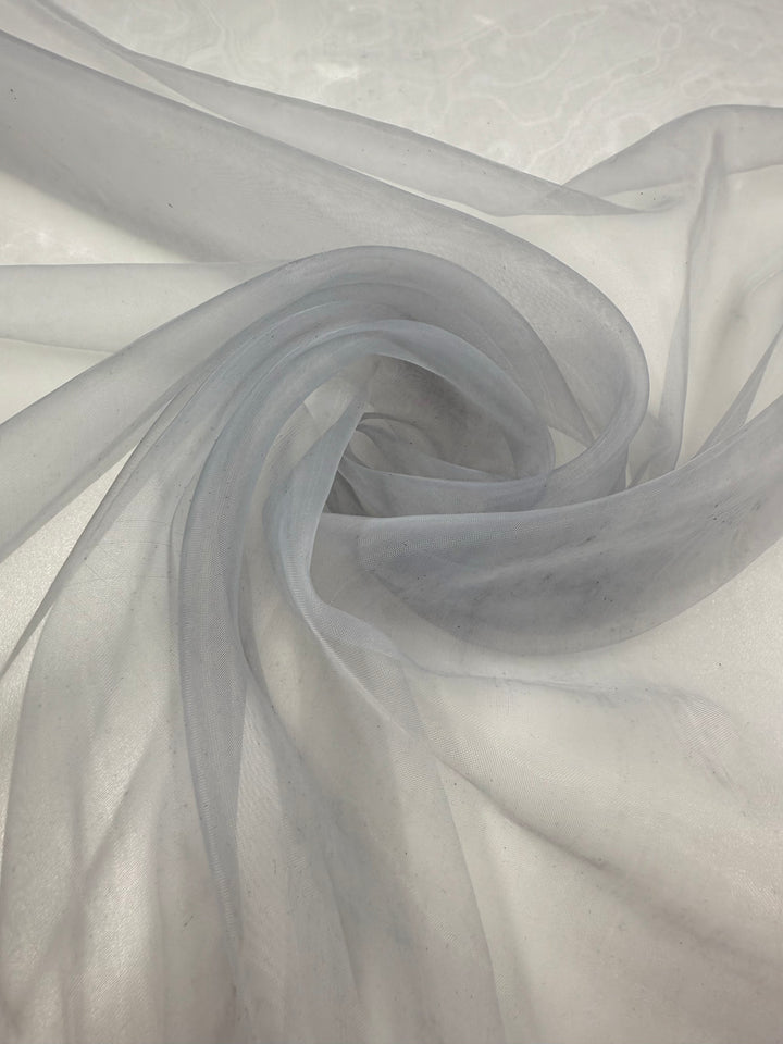 A close-up of Super Cheap Fabrics' Organza - Illusion Blue - 150cm shows sheer fabric swirling gently on white, creating delicate folds and a translucent, ethereal look.