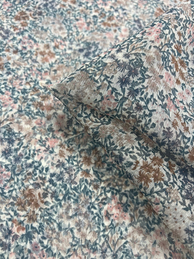 A close-up of a folded section of the Pure Printed Silk Chiffon - Soft Flowers from Super Cheap Fabrics reveals a dense floral pattern. The design features small, detailed flowers in shades of pink, blue, and brown set against a light background. This 132cm wide chiffon fabric appears soft and lightweight, epitomizing high-end fashion luxury fabric.