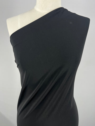 A mannequin draped in a one-shoulder dress made from ITY Knit - Black - 150cm fabric by Super Cheap Fabrics is shown against a plain white background. Crafted from black interlock twist yarn fabric, the dress has a smooth texture and snug fit, highlighting its elegant and minimalist design. The image focuses on the torso and part of the medium weight fabric dress.