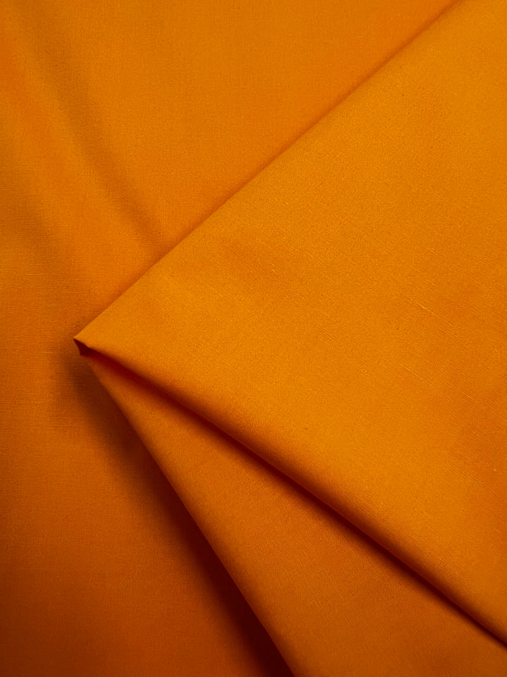 A close-up of Super Cheap Fabrics' Plain Cotton - Zinnia (147cm), neatly folded to display its bright orange color and smooth, lightweight 100% cotton texture—ideal for vibrant, airy creations.