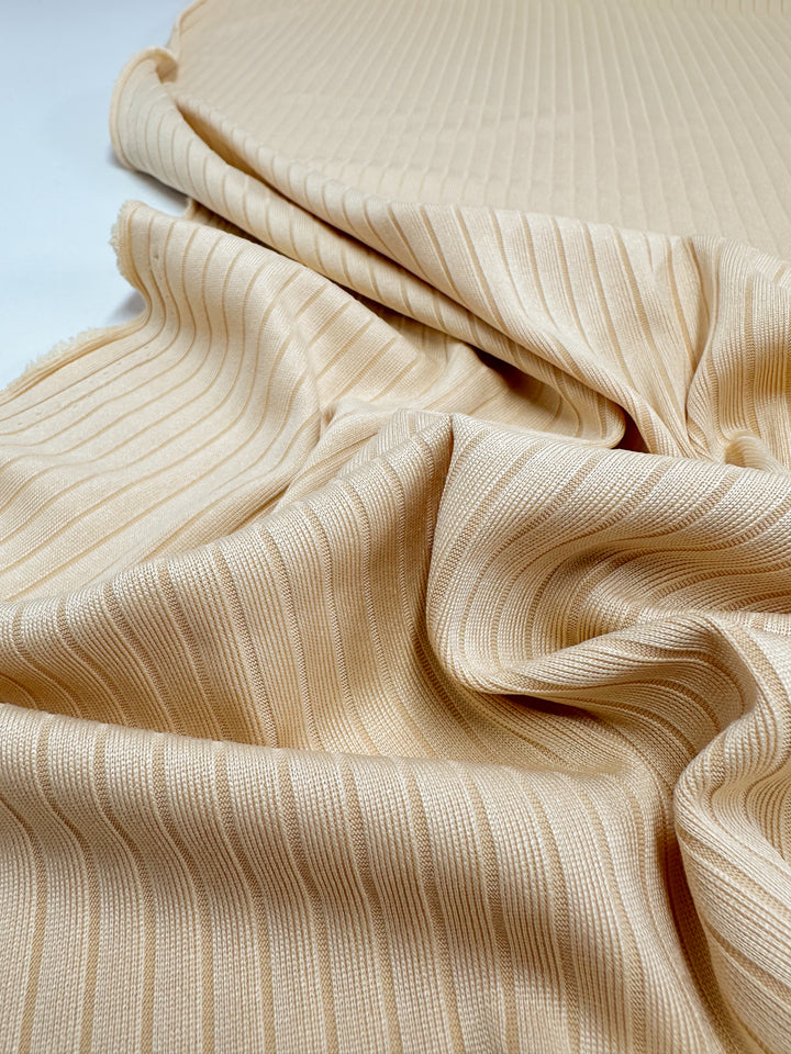 The Apricot Gelato Rib Knit from Super Cheap Fabrics, measuring 170cm, features a soft beige ribbed fabric draped in folds. It has a subtle sheen with vertical lines that add depth and flow to its textured surface.
