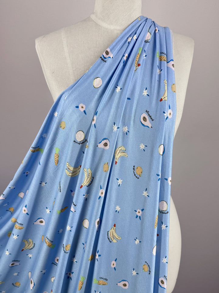 The Super Cheap Fabrics Printed Cotton Lycra - Fruity (150cm) draped over a mannequin features a playful avocado, banana, and egg pattern on a light blue bell background. Made from cotton spandex, this fabric is both stylish and comfortable with its whimsical touch.