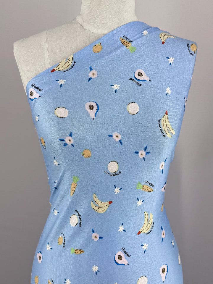 Product: Printed Cotton Lycra - Fruity - 150cm by Super Cheap Fabrics. A playful, tropical design featuring bananas, pineapples, coconuts, and star motifs on blue bell fabric. Crafted from lightweight cotton spandex and elegantly draped over a mannequin.