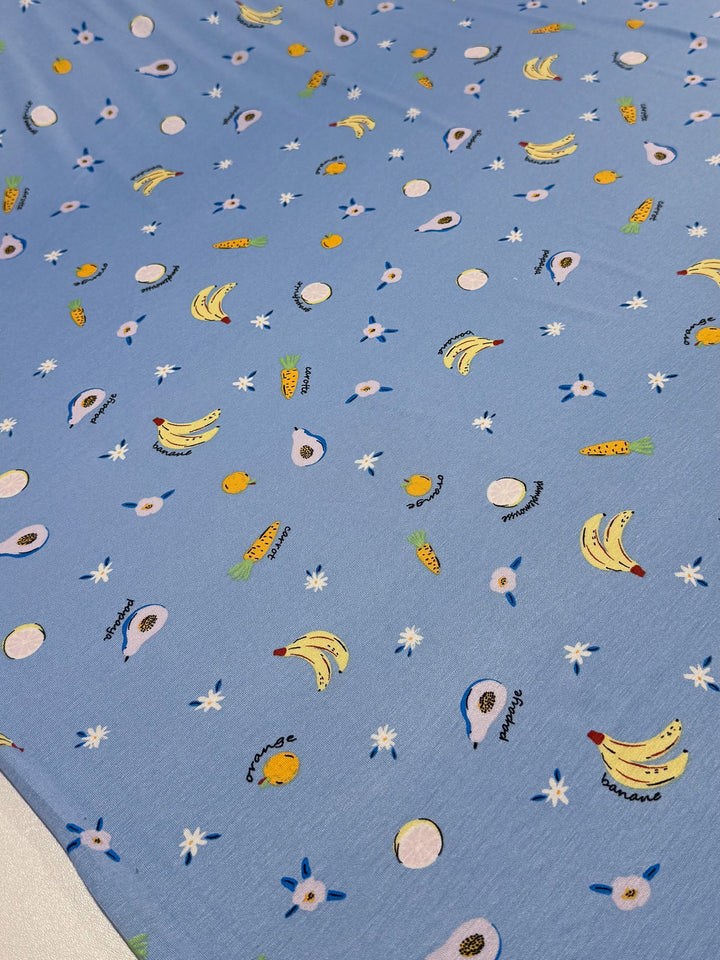 The "Printed Cotton Lycra - Fruity" fabric by Super Cheap Fabrics features a scattered print of bananas, avocados, pineapples, and kiwis with playful labels on bluebell-colored cotton spandex. White star-like shapes are interspersed throughout this delightful medium-weight 150cm wide design.