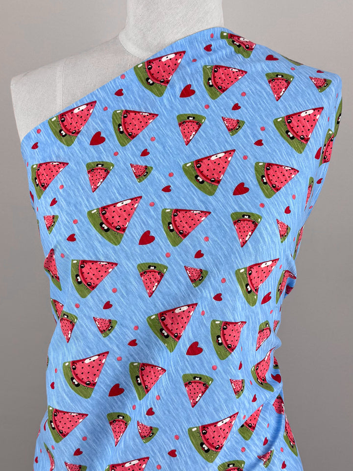 The Super Cheap Fabrics' Printed Jersey - Sandia is draped on a mannequin to showcase its playful blue print of red watermelon slices and small hearts, highlighting the vibrant, multi-color design on this 145cm medium weight fabric.