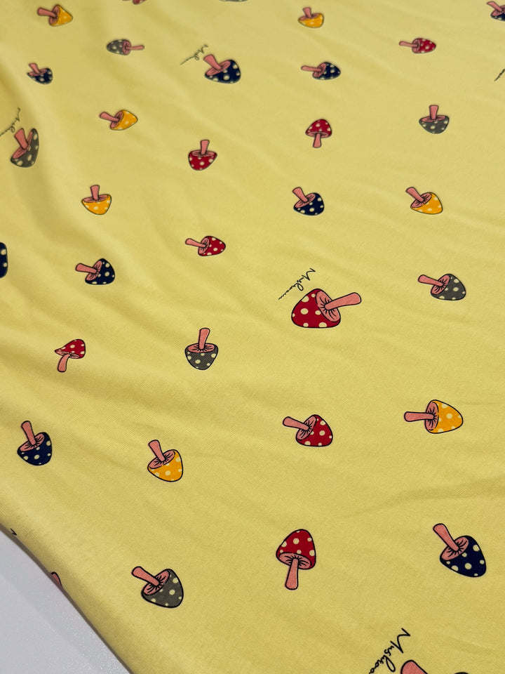 The Printed Jersey - Shroom by Super Cheap Fabrics is a yellow medium-weight fabric with a playful pattern of red, yellow, and black mushrooms, some dotted. "Mushroom" text appears near select mushrooms. The fabric offers slight two-way stretch for comfort and measures 160cm.
