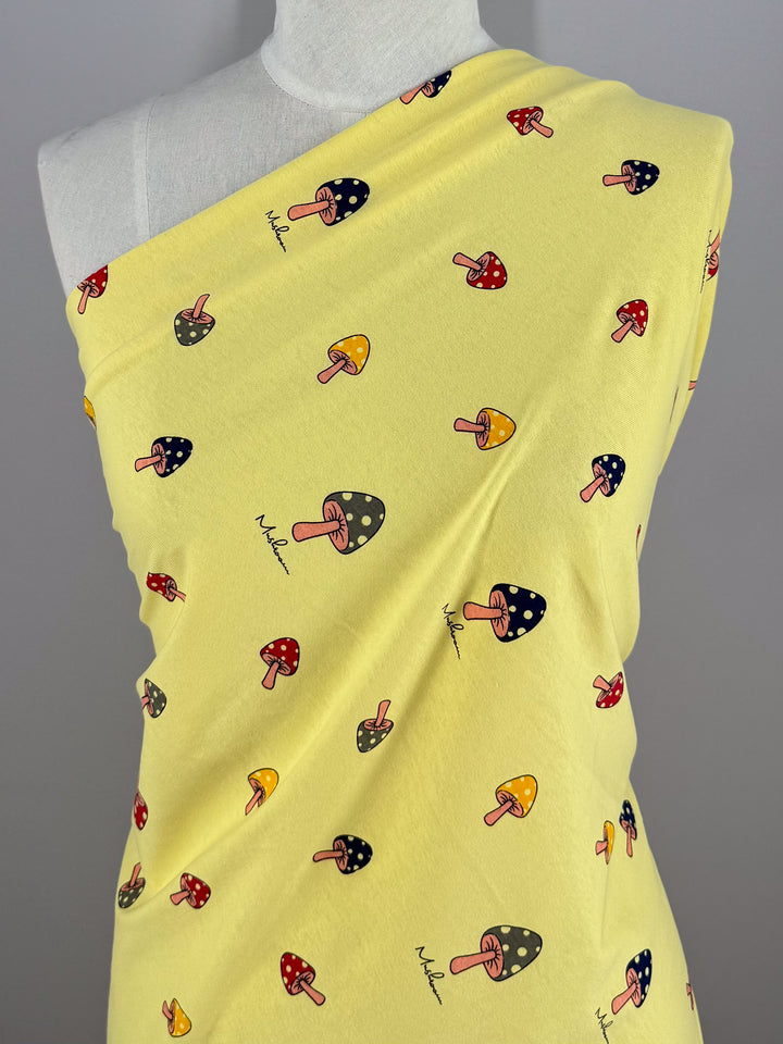 Super Cheap Fabrics' Printed Jersey - Shroom (160cm) features a yellow base with vibrant red, yellow, and blue mushroom motifs. Medium weight and elegantly draping over mannequins on a gray background, it offers a gentle two-way stretch for versatility.