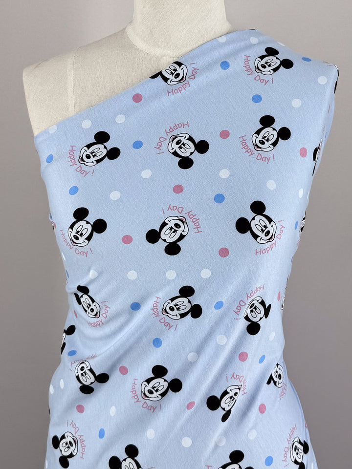 The "Cheers Ears" Printed Jersey by Super Cheap Fabrics features a cheerful pattern of cartoon mouse faces with "Happy Day!" in pink and red, draped over a mannequin. This medium-weight fabric, 150cm wide, is adorned with white, pink, and blue polka dots scattered throughout.