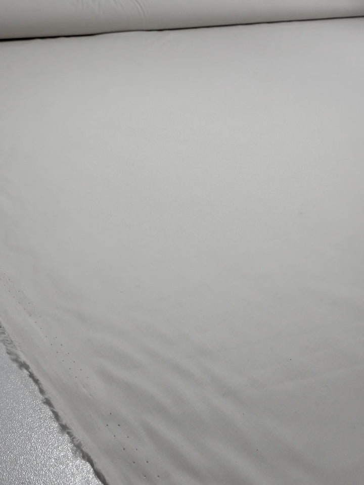 A close-up of Super Cheap Fabrics' Cotton Voile - Gardenia (150cm) shows a large, smooth roll of plain white fabric spread flat, with its edge visible in the lower left corner.