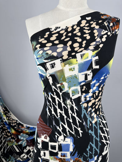 A mannequin draped in Super Cheap Fabrics' vibrant Printed Lycra - Tonal - 150cm with an abstract print. The design features a mix of geometric shapes, houses, and splashes of blue, black, white, and green. The background is a neutral grey, highlighting the fabric's intricate pattern.