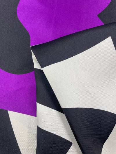 A close-up image of the Silk Twill Panel - Crop - 145cm by Super Cheap Fabrics, featuring bold, abstract shapes in vibrant purple, white, and black. The luxurious material is draped in folds, showcasing the smooth texture and striking color contrast.