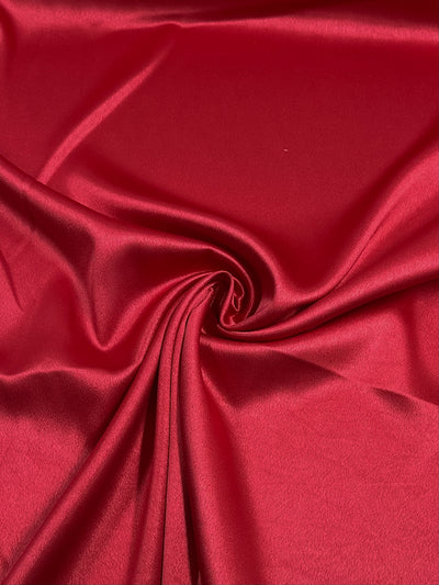 A close-up image of the Satin Back Crepe in Crimson by Super Cheap Fabrics, showcasing its smooth and glossy texture with subtle folds and a gentle swirl at the center. The material's reflective surface highlights its luxurious, silky appearance.