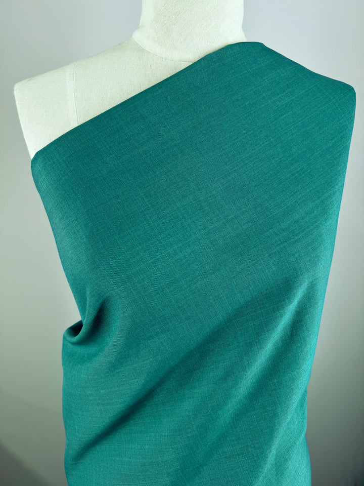 A mannequin displays Super Cheap Fabrics' Twill Suiting - Ivy - 150cm, draped over one shoulder to showcase its smooth, teal-green hue and diagonal twill weave against a plain light background.