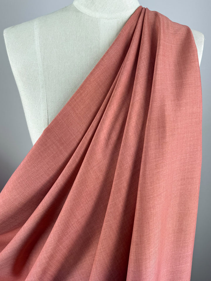 Super Cheap Fabrics’ Twill Suiting - Terra Cotta (150cm), a 100% polyester fabric with a pinkish-coral hue and diagonal weave, is draped over a cream dress form, showcasing its soft, elegant folds against a neutral background.