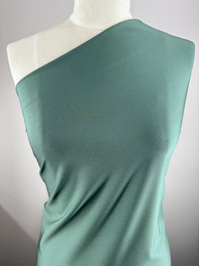 A dress form displays Super Cheap Fabrics' Nylon Lycra - Bistro Green (154cm), draped diagonally to cover one shoulder. The smooth four-way stretch fabric creates a sleek silhouette against a light background.