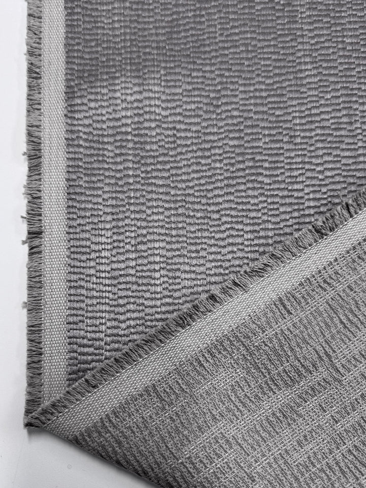 A close-up shows Super Cheap Fabrics' Upholstery Jacquard - Oscar Dusk (142cm), a gray textured heavyweight fabric with fringed edges, folded to display both front and back sides.