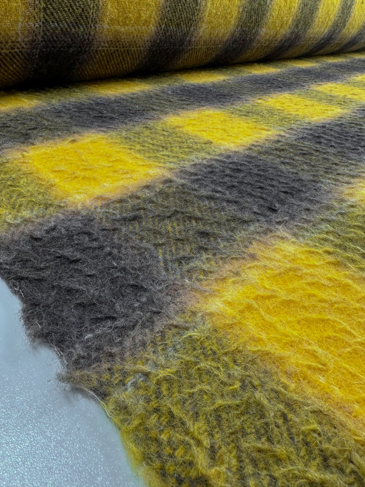 Close-up of a piece of Virgin Wool - Spruzzo - 145cm from Super Cheap Fabrics, showcasing its soft, fuzzy texture. This heavy-weight wool fabric features an inviting plaid pattern with alternating yellow and dark gray squares, giving it a cozy and warm appearance. Partially unfurled to reveal the intricate pattern up close, it's ideal for crafting overcoats.