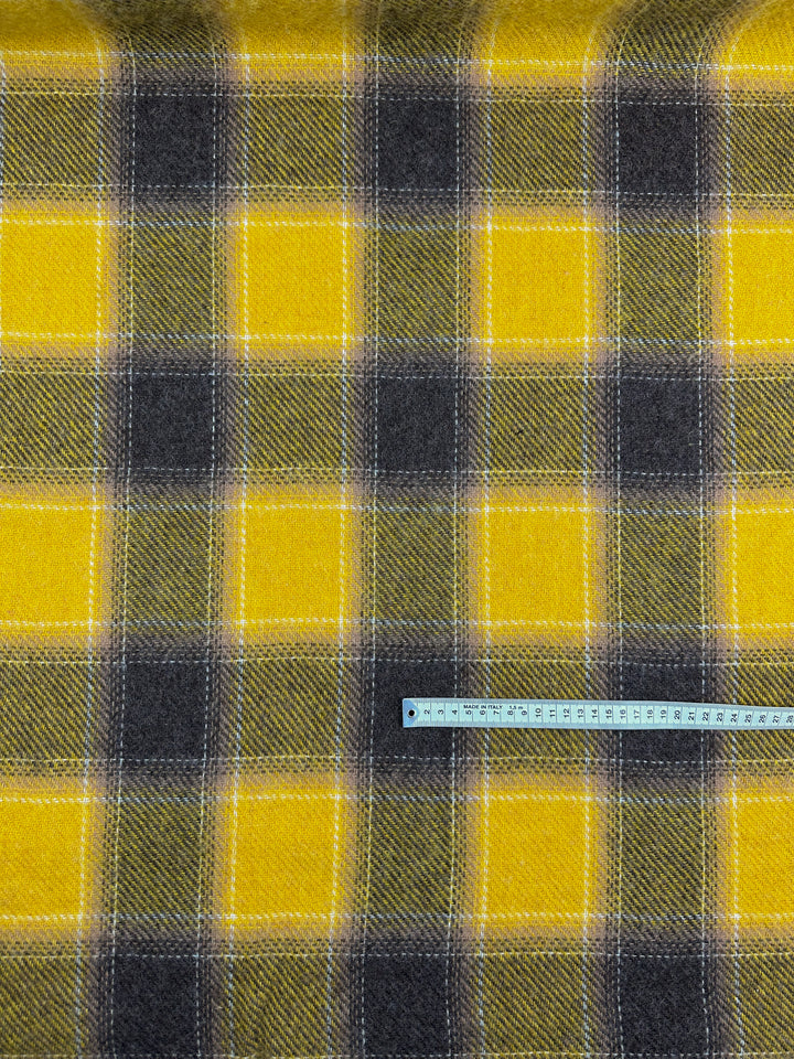 A fabric sample of "Virgin Wool - Spruzzo" by Super Cheap Fabrics, featuring a heavy-weight yellow and gray tartan pattern with large squares. A blue measuring tape stretched across the bottom in centimeters indicates the scale of this 145cm wide virgin wool pattern, ideal for overcoats.