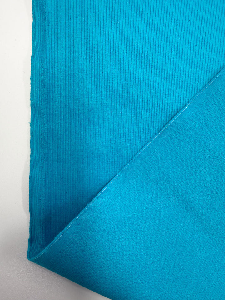 A close-up of Super Cheap Fabrics’ Pin Wale Corduroy - Blue Jewel (147cm) shows its ribbed and smooth textures, folded at one corner against a white background.