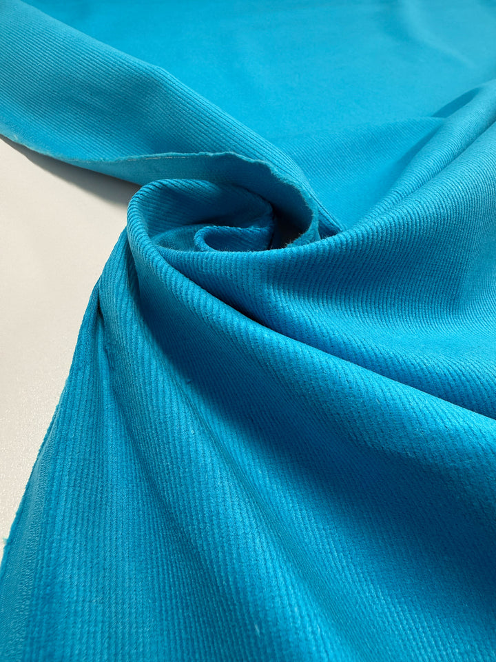 A close-up of Super Cheap Fabrics' Pin Wale Corduroy in Blue Jewel (147cm), partially folded and twisted on a white surface to showcase its ribbed texture and soft drape.