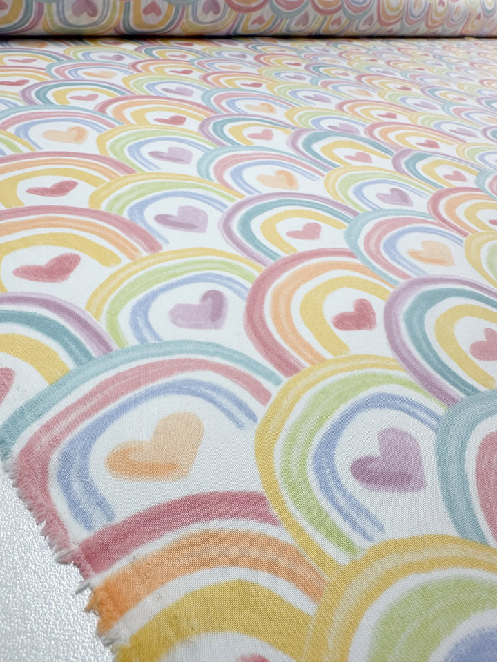 Close-up of the Unity Printed Delsutered Satin from Super Cheap Fabrics showcases overlapping rainbows with heart centers. The pastel hues in pink, orange, yellow, green, and blue form a whimsical design on this lightweight 155cm fabric reminiscent of printed satins.