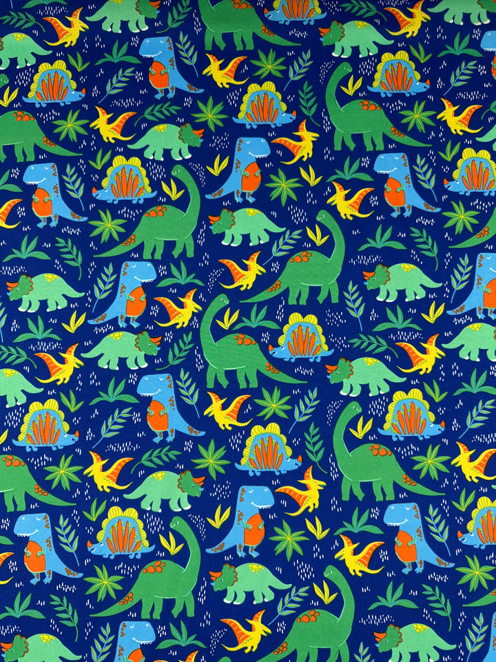The Dino Jungle delustered satin from Super Cheap Fabrics features a dark blue background adorned with triceratops, pterodactyls, and stegosauruses in vibrant green, orange, and blue among lush foliage for an elegant print.