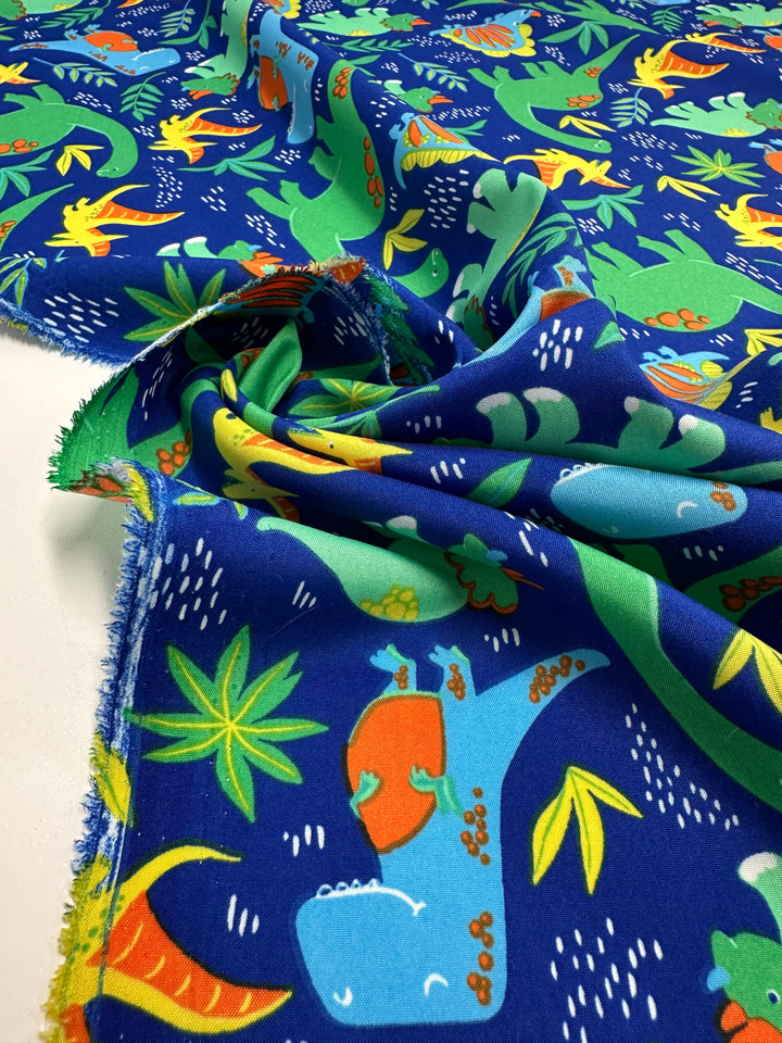The Super Cheap Fabrics' Printed Delustered Satin - Dino Jungle - 150cm features a playful dinosaur and plant pattern on a vibrant blue background, with dinosaurs in green, orange, and teal shades. The fabric elegantly spreads out with slight gathering in the center.