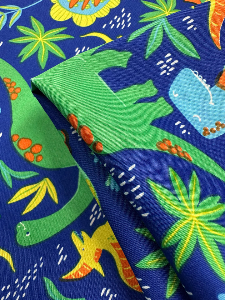 The Printed Delustered Satin - Dino Jungle from Super Cheap Fabrics features playful green, blue, and yellow dinosaurs mixed with green and yellow tropical leaves on a pristine blue background. Width: 150cm.