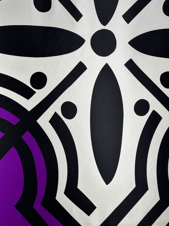 The Silk Twill Panel - Crop - 145cm from Super Cheap Fabrics showcases a luxurious geometric design featuring bold, black shapes such as ovals, circles, and lines symmetrically arranged on a white background. This abstract pattern also includes a distinctive purple section in the bottom left corner.