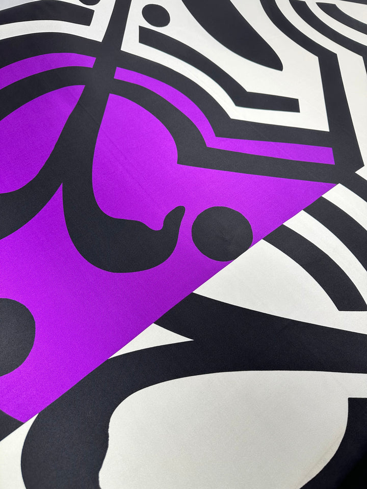 A close-up of the "Silk Twill Panel - Crop - 145cm" from Super Cheap Fabrics showcases an abstract geometric design with bold black lines and shapes on a white background, highlighted by a vibrant purple section. The pattern forms intriguing shapes and curves, reminiscent of silk twill fabric, creating a striking visual contrast.