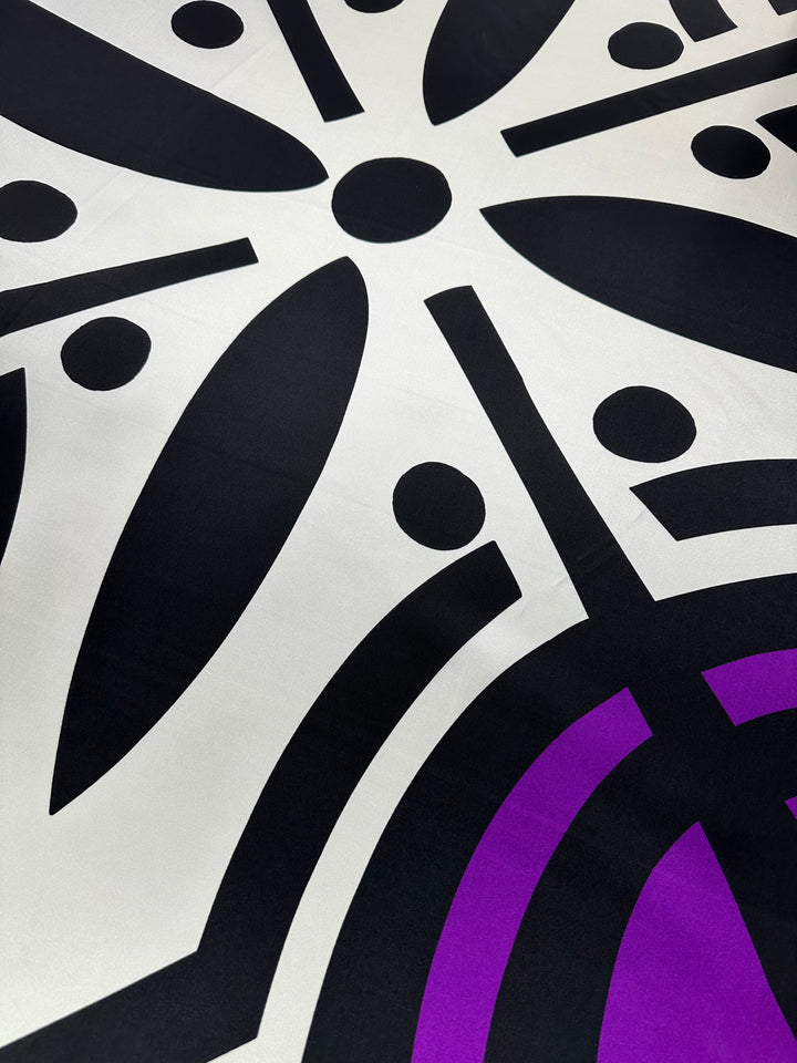 A close-up view of the Silk Twill Panel - Crop - 145cm by Super Cheap Fabrics, showcasing a geometric design with black and purple shapes on a white background. The pattern, reminiscent of intricate silk fibers, features symmetrically arranged circular and leaf-like forms.