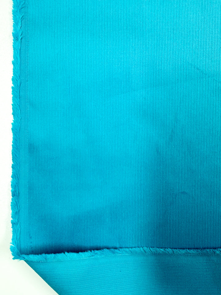 A close-up image of a piece of Micro Wale Corduroy in the Scuba color, featuring a textured surface with visible fibers along the edges. The fabric is turned up at the corner, showing both the front and back sides, making it perfect for winter clothing or stylish home decor. This high-quality fabric from Super Cheap Fabrics measures 140cm in width.