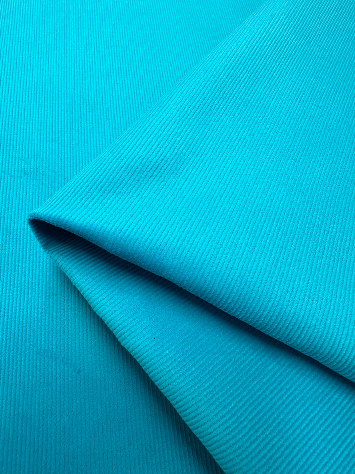 Close-up of a folded piece of Super Cheap Fabrics' Micro Wale Corduroy in Scuba. The material appears smooth and soft, with a subtle ribbed texture reminiscent of corduroy fabric. The folds create gentle shadows and highlights, adding depth. This vibrant color can bring a fresh touch to winter clothing or home decor.