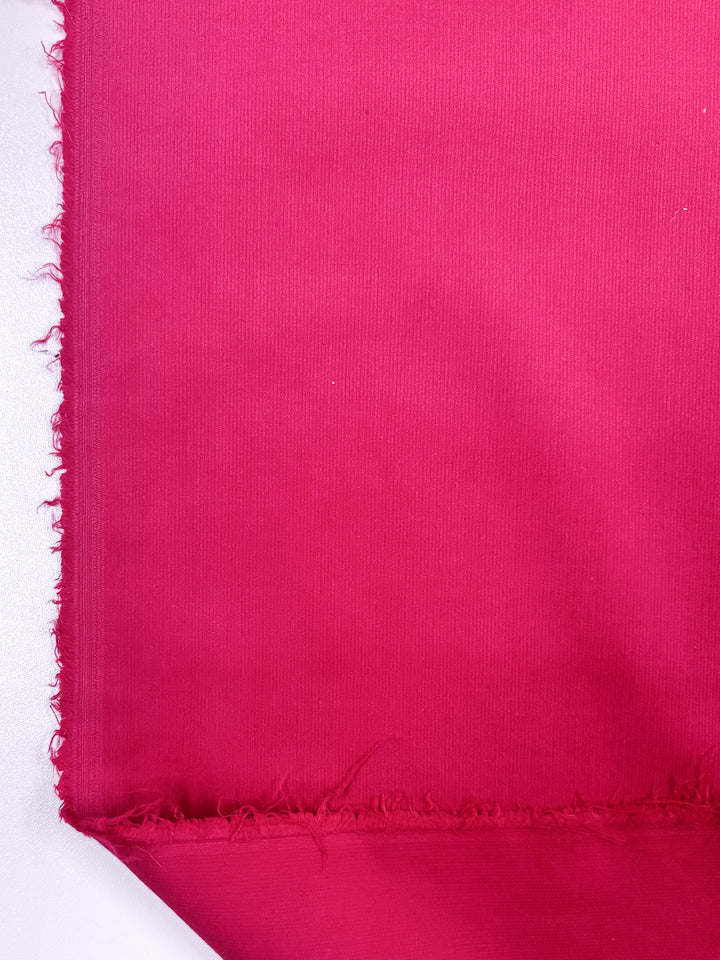 Close-up of a piece of Micro Wale Corduroy - Raspberry Sorbet from Super Cheap Fabrics, showcasing its vibrant hue on a white background. The textured weave and frayed edge on the left side suggest it could be perfect for winter clothing or stylish home decor items.