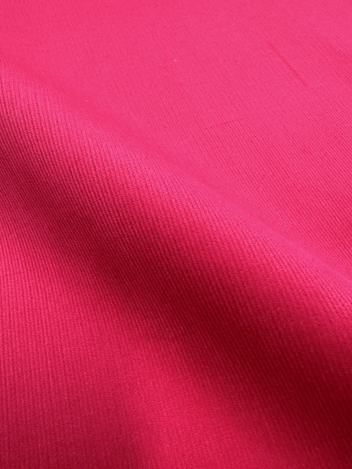 A close-up view of the vibrant Raspberry Sorbet Micro Wale Corduroy fabric by Super Cheap Fabrics, highlighting its texture and weave. The material has a smooth surface with subtle diagonal lines, suggesting a classic twill pattern. Ideal for home decor items or cozy winter clothing, the slightly curved fabric creates gentle shadows and highlights across its 147cm width.