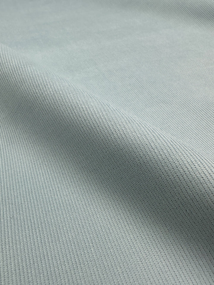 A close-up view of the Spa Blue Micro Wale Corduroy from Super Cheap Fabrics reveals its light grayish-blue hue and subtle diagonal ribbed texture. The 145cm wide material appears smooth and soft, with gentle shadows accentuating the ridges and folds, making it an ideal choice for home decor or cozy winter clothing.