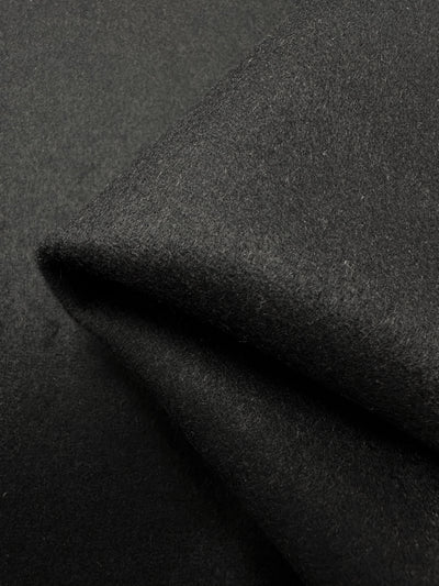 Image of two overlapping pieces of Super Cheap Fabrics' Wool Cashmere - New Black - 150cm. The dark grey felt fabric resembles heavy weight wool fabric, with a texture that appears soft and slightly fuzzy, creating a smooth and plush surface. Ideal for outer coats, the edges of the black fabric are shown in the two overlapping pieces.