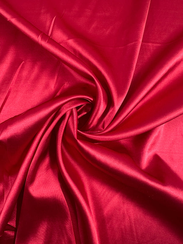 The Scarlet Smile from Super Cheap Fabrics is a vibrant red satin, expertly gathered and twisted to highlight its smooth, shimmering texture. The deluxe material's central swirl elegantly reflects light on its silky surface.