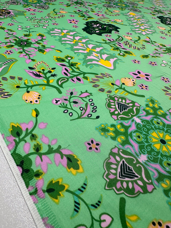 A close-up of Cotton Sateen - Lime Mosaic (145cm) by Super Cheap Fabrics, showing a vibrant floral print with pink, yellow, and dark green on a light green background. The fabric edge is visible, ideal for household décor projects.